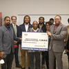 A group of 12 people pose for a picture while holding a large novelty check.