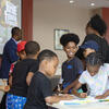 Children stand around a table while coloring and writing on paper.